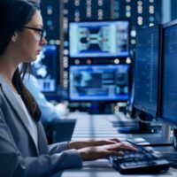 Female Engineer Controller Observes Working of the System. In the Background People Working and Monitors Show Various Information.